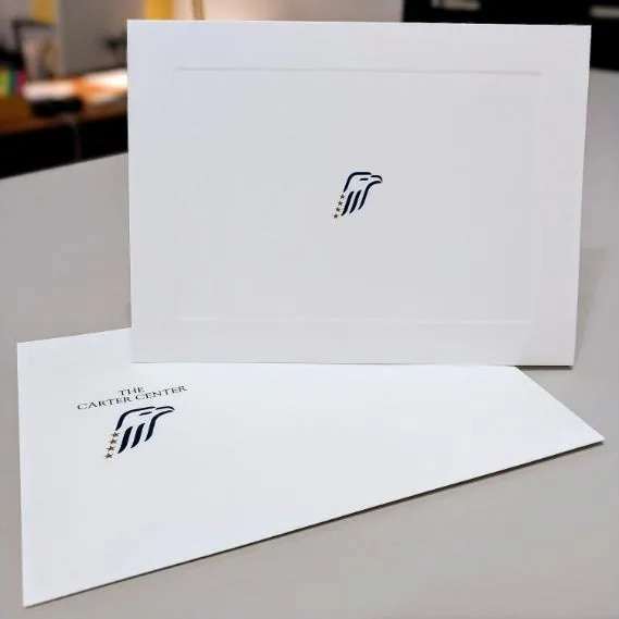 A white note card with a blind-embossed panel border and the Carter Center eagle mark, beside its engraved envelope, photographed on the Brookwood Printing worktop
