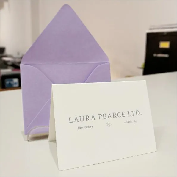 A folded ecru note card standing in front of an opened lilac-lined envelope